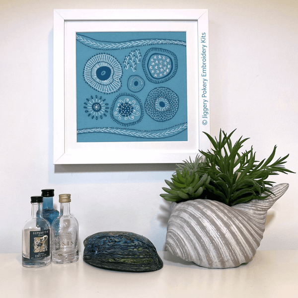 Abstract embroidery kit shown completed and hanging on cream wall with miniature gin bottles and plant for scale