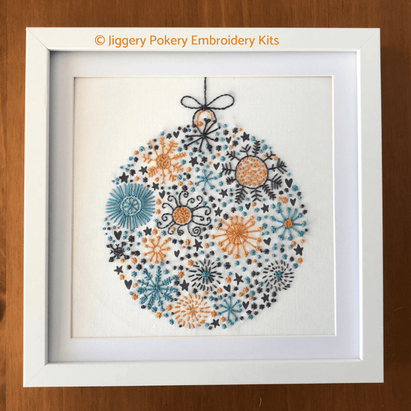 Framed festive ball Christmas hand embroidery pattern in white mount and frame, shown on wooden background
