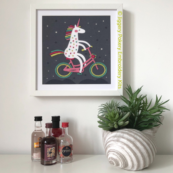 Framed unicorn embroidery hanging on wall with miniature gin bottles and plant for scale