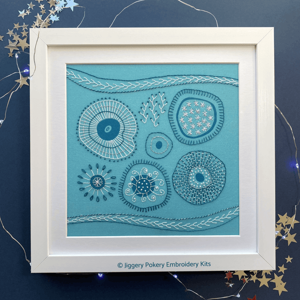 Abstract embroidery kit mounted in square white frame, shown with lights and stars on a navy blue background