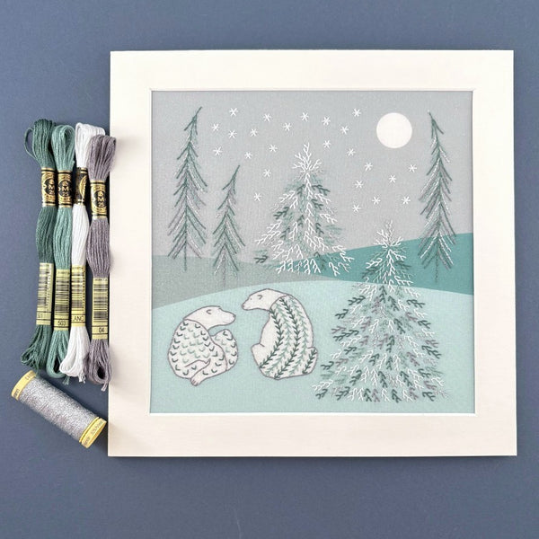 Framed winter scene with trees and polar bears, shown on a blue background with DMC embroidery threads in white, grey, green with Gutermann silver metallic effect thread.