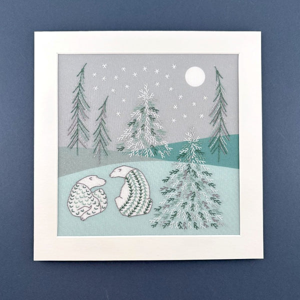 Bears embroidery of two decorated polar bears on a green and grey background with frozen trees. Placed in a square white mount on a dark blue background.