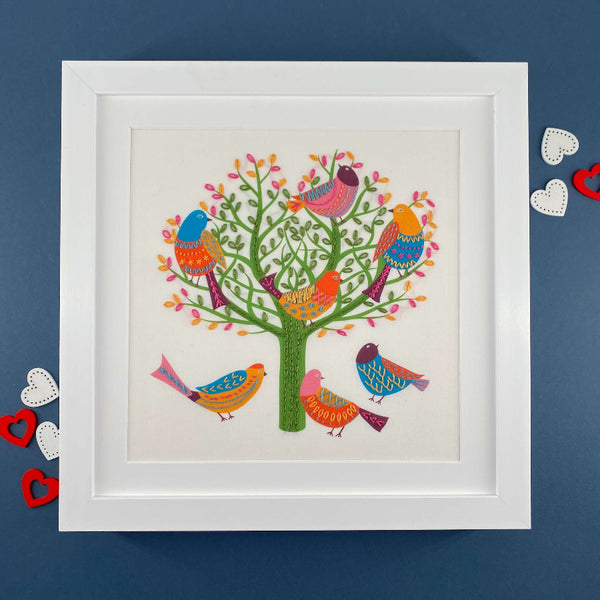 Birds in a tree embroidery pattern shown in a square white frame with coloured hearts on a navy blue background