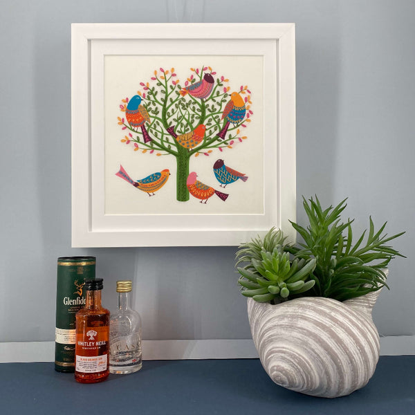 Bird embroidery mounted in a square white frame hanging on a grey wall, shown with mini gin bottles for scale.