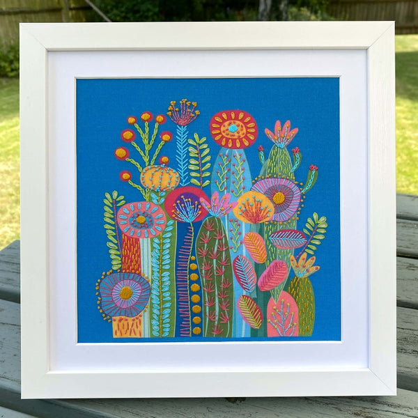 Cactus embroidery design on blue fabric shown in a square white frame standing outdoors on a garden table