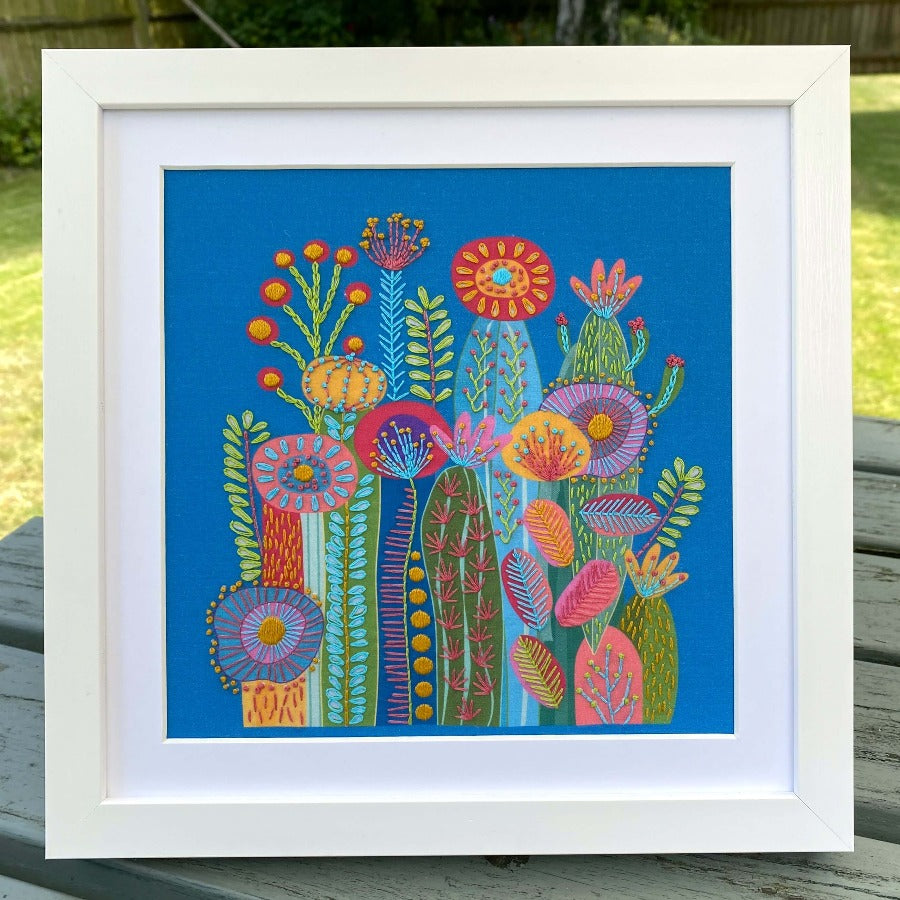Cactus embroidery kit completed on blue fabric mounted in a square white frame. Photo taken in a sunny garden.