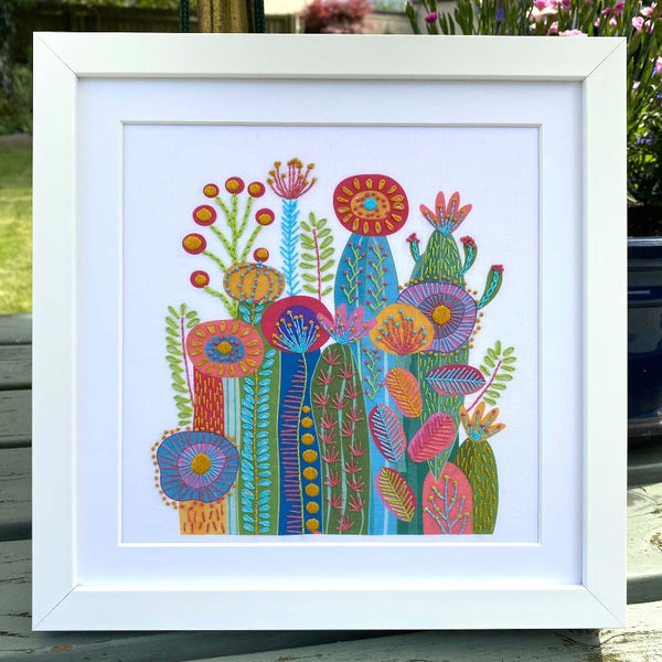 Cactus embroidery design on white fabric shown in a square white frame standing outdoors on a garden table