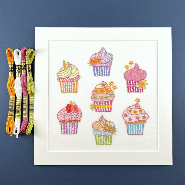 Cupcakes embroidery in white mount shown with DMC threads in gold, white, pink and green.