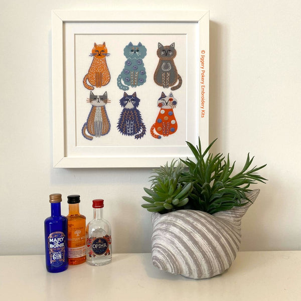 Cat embroidery in a square white frame hanging on a cream wall with miniature gin bottles and a plant for scale