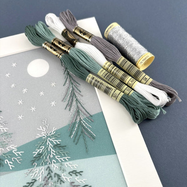 Set of DMC embroidery threads in white, green and grey with a reel of Gutermann metallic effect thread shown with a close-up of two trees in the bear embroidery kit. 