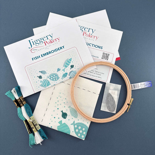 Contents of the fish embroidery kit include a wooden embroidery hoop, needles and a needle threader in a paper bag, the fish design printed on fabric, 3 skeins of DMC thread plus instructions. Shown on a navy blue background