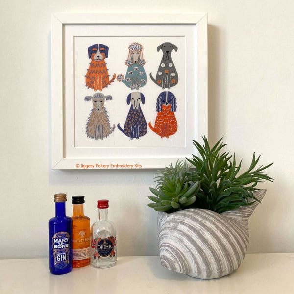 Dog embroidery kit in a square white frame hanging on a cream wall with a plant for scale