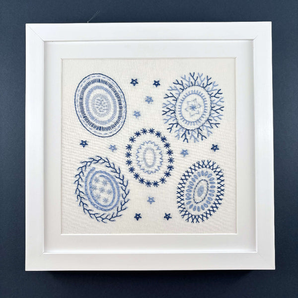 Learn embroidery sampler kit mounted in a square white frame shown on a navy blue background.
