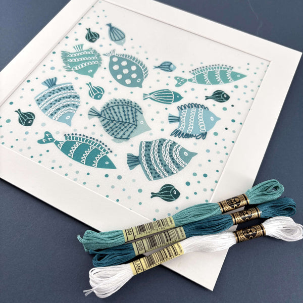 Fish embroidery kit in a square white mount with 3 skeins of teal, turquoise and white DMC threads. Shown on a navy blue background.