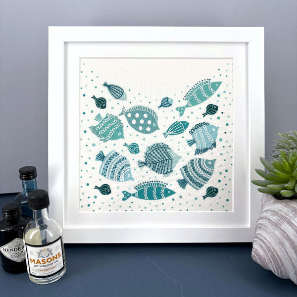 Fish embroidery kit mounted in a square white frame. Shown standing on a dark blue shelf against a grey wall. Mini gin bottles and a plant included for scale.