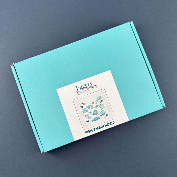 Turquoise box containing the fish embroidery kit. The box has a Jiggery Pokery label showing the design and a list of contents. Shown on a dark blue background