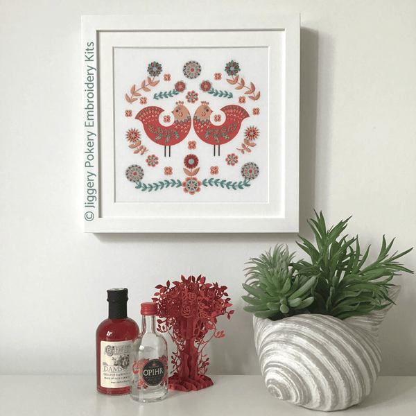 Framed embroidery of folk art birds shown with miniature bottles and plant for scale