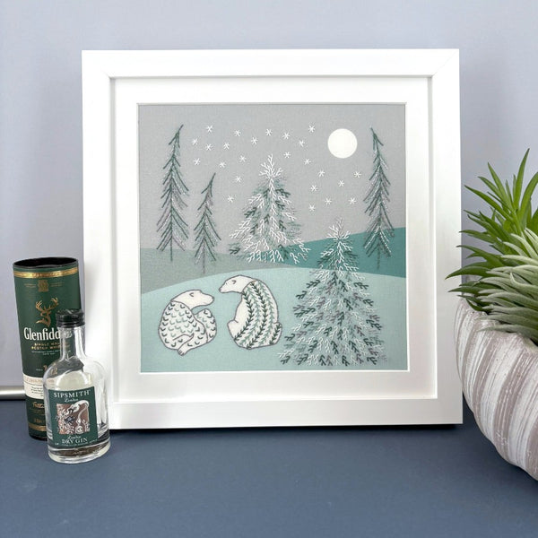 Framed embroidery of two polar bears in a snowy landscape with trees, next to mini bottles and a plant for scale. Shown standing on a blue shelf.