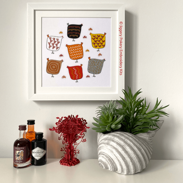 Chicken embroidery in square white frame hanging on wall with miniature gin bottles and plant for scale
