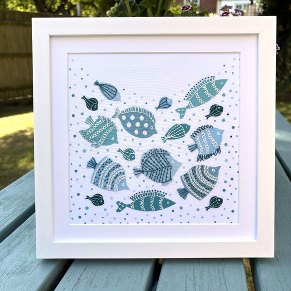 Framed fish embroidery showing a shoal of blue and green fish swimming through bubbles. Mounted in a square white frame, shown standing on a garden table.