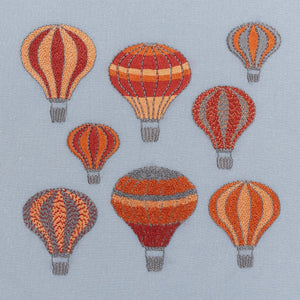 Embroidery kit showing a group of hot air balloons stitched in ruby red, apricot, orange and grey drifting on a dark grey sky