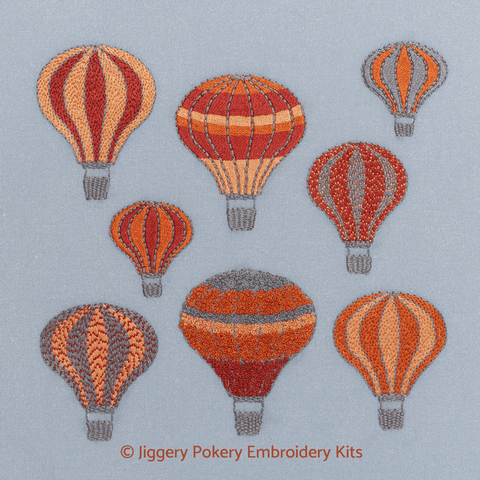 Embroidery kit showing a group of hot air balloons in ruby red, apricot, orange and grey drifting on a dark grey sky