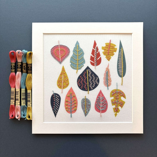 Jiggery Pokery leaf embroidery shown in white mount with dark pink, pale pink, blue and mustard DMC threads on a dark blue background
