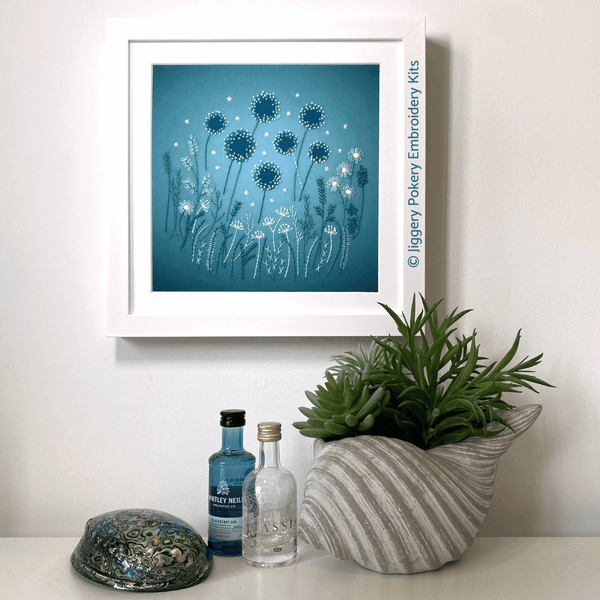 Modern embroidery flowers kit in a square white frame hanging on a wall with a plant and mini gin bottles for scale.