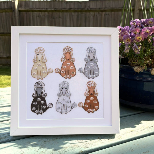 Poodles embroidery mounted and in a square white frame. Shown standing on an outdoors table next to a plant