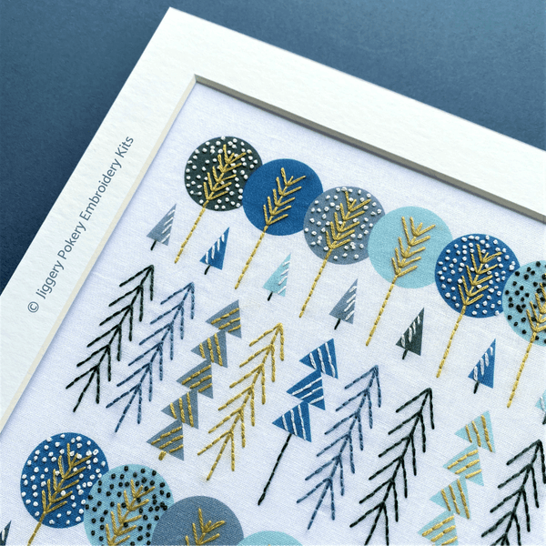 Close-up of simple trees embroidery design in white mount, stitched in gold, white, dark green and blue.
