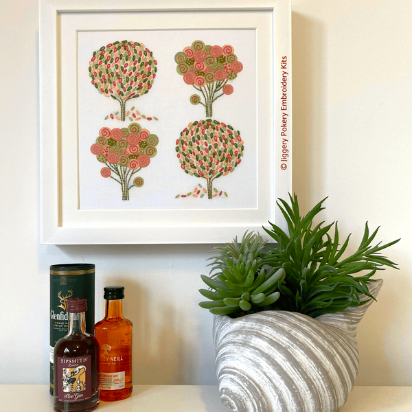 Spring embroidery kit in square white frame hanging on a wall. Shown with mini gin bottles and a plant for scale.