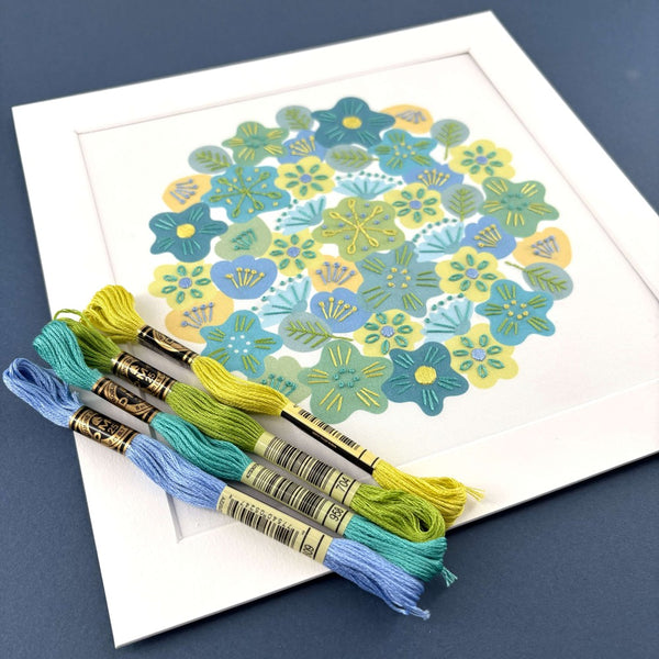 Spring flowers embroidery in a white mount on a dark blue background. Shown with DMC threads in yellow, green, turquoise and blue
