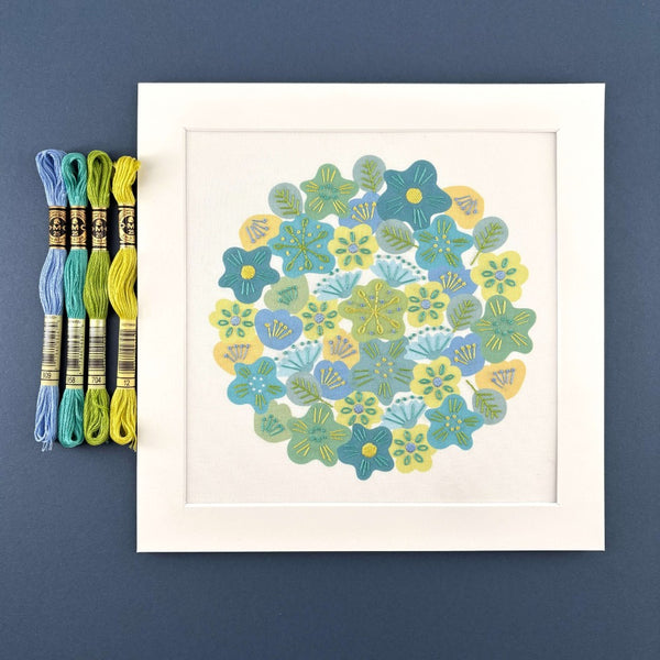 Spring flowers embroidery in a square white mount, shown with 4 skeins of DMC thread in yellow, grass green, turquoise and blue on a navy blue background