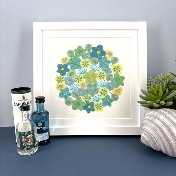 Spring flowers embroidery in a square white frame. Shown standing on a dark blue shelf with mini gin bottles for scale and a plant.