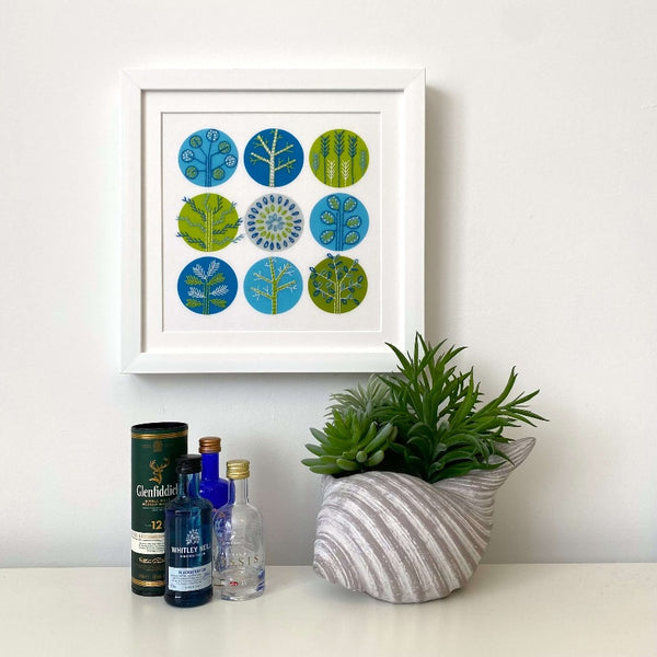 Blue and green trees embroidery pattern shown mounted in a square white frame hanging on a wall. Mini gin bottles and a plant shown for scale.