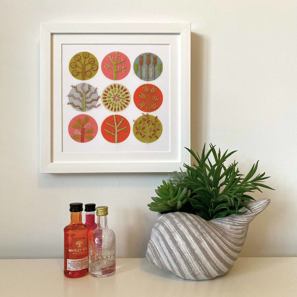 Pink and orange Scandi style trees embroidery shown mounted in a square white frame hanging on a wall. Shown with mini gin bottle and a plant for scale.