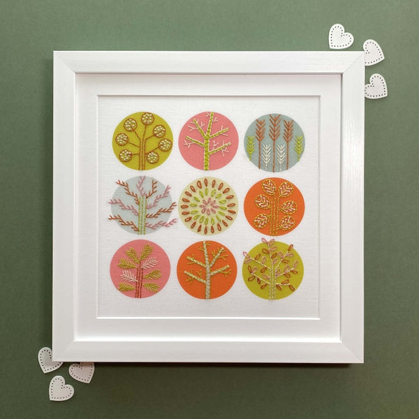 Orange and pink trees embroidery pattern mounted in a square white frame, shown on a green background with hearts