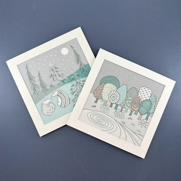 Winter trees embroidery pattern with bears embroidery pattern, both in white square mounts. Shown on a navy blue background.