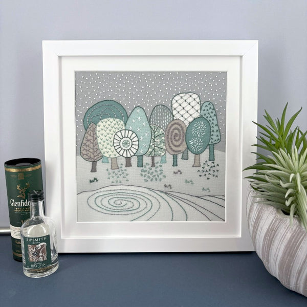 Winter landscape embroidery mounted in a square white frame. Shown on a shelf against a grey wall with mini drinks bottles and plant for scale.