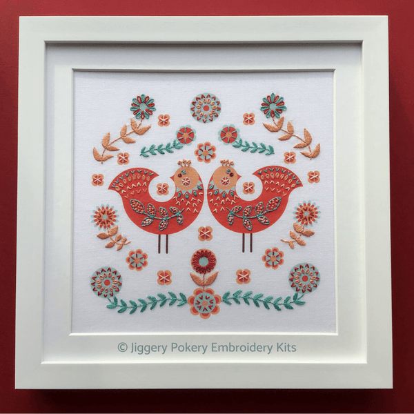 Framed folk embroidery kit showing two red birds surrounded by decorations in turquoise blue and orange stitching
