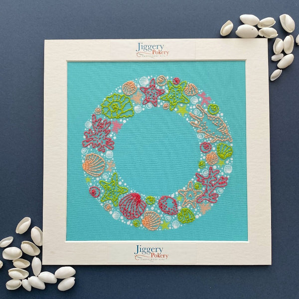 Seaside embroidery wreath pattern in white mount on dark blue background shown with seashells
