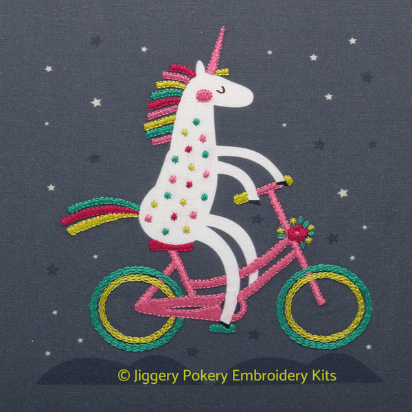 Unicorn embroidery kit showing a brightly coloured unicorn riding a bicycle against a dark grey background