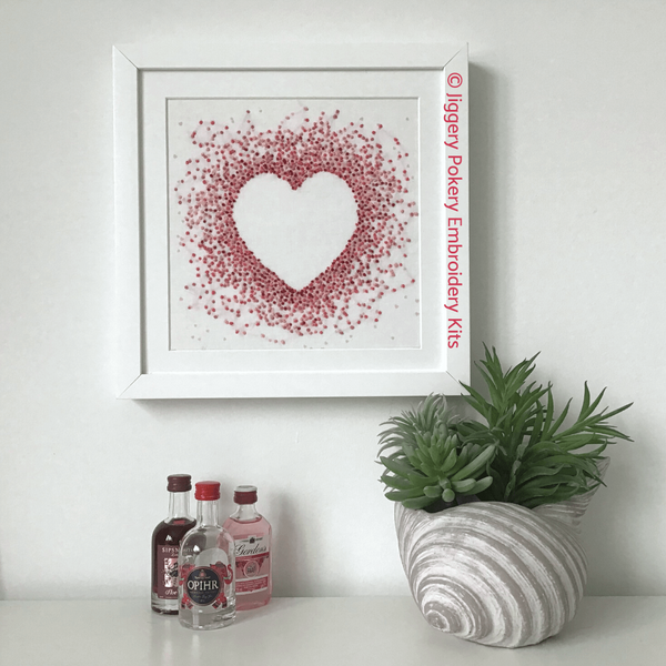 Pink hearts embroidery kit by Jiggery Pokery framed and hanging on wall, shown with miniature gin bottle for scale