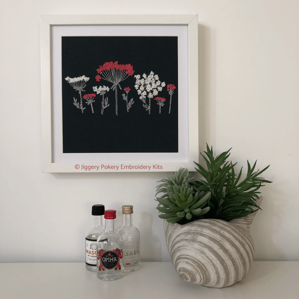 Wildflower embroidery pattern framed and hanging on a cream wall with miniature bottles and a plant for scalewall